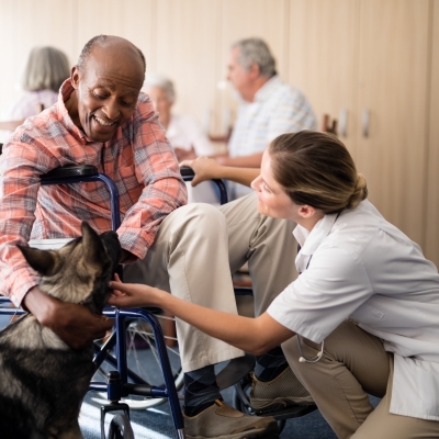 Cheerful female doctor kneeling by disabled senior man stroking puppy at retirement home