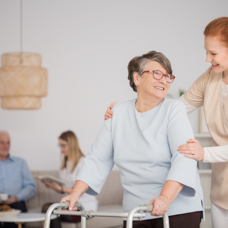 Happy nurse assisting a senior woman with a walker and another nurse reading a book to a pensioner in the background